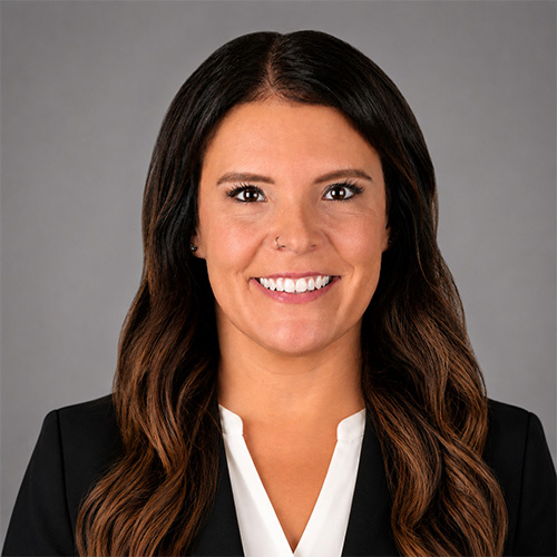 Portrait of a smiling woman with long dark hair, wearing a black blazer and white blouse against a gray background.